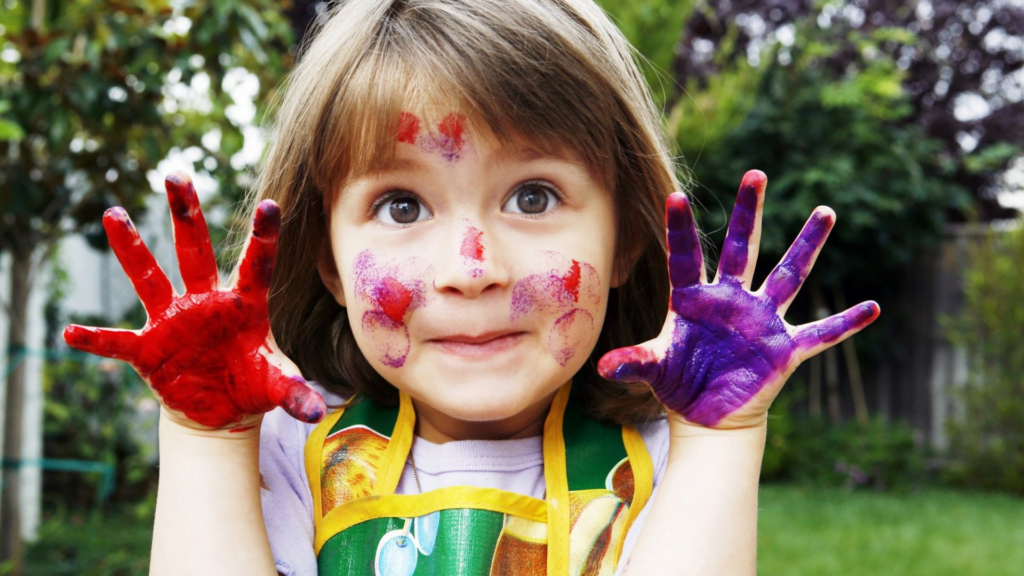 Es una niña, con una mano pintada de color rojo y otra mano de color púrpura. Tiene manchas de pintura en la cara.