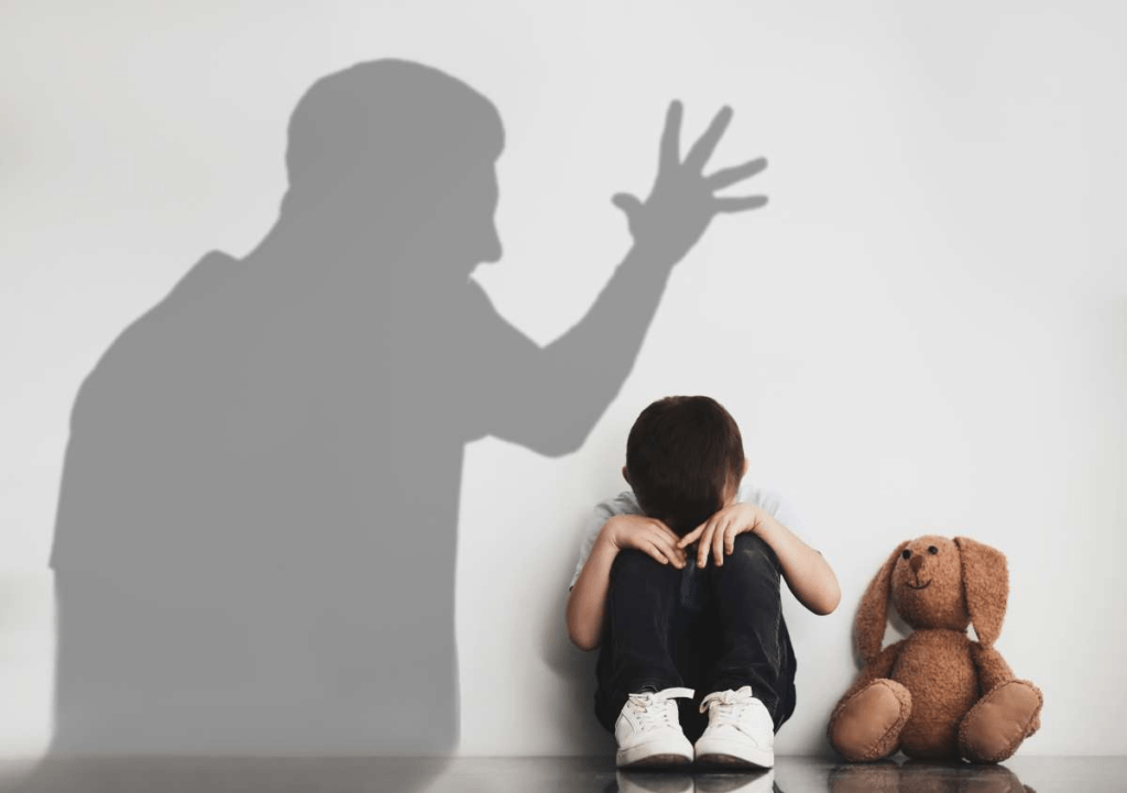 Imagen con una sombra que pertenece a un hombre adulto, que está amenazando con su mano a un niño pequeño, que está sentado con un oso de peluche.